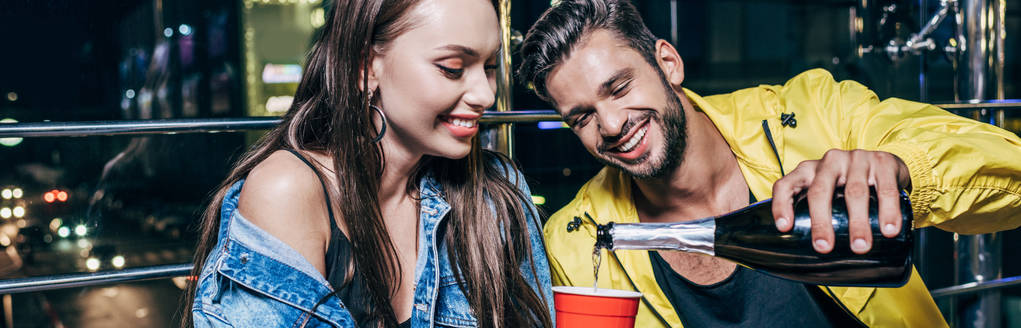 Panoramaaufnahme eines gutaussehenden und lächelnden Freundes, der in der Nacht Champagner in Plastikbecher gießt  - Foto, Bild