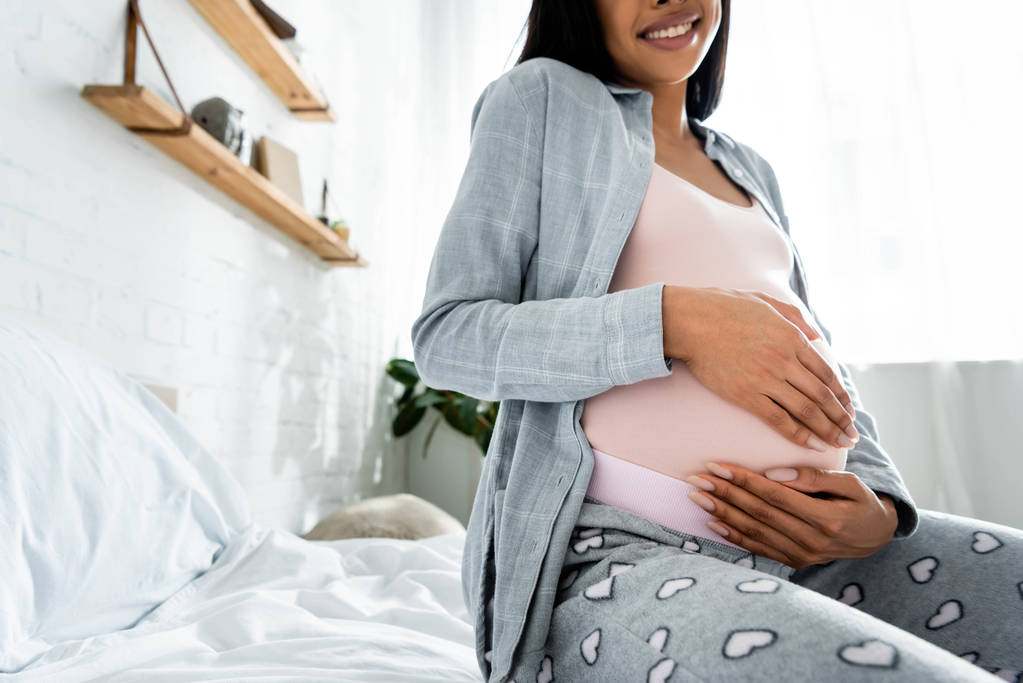 abgeschnittene Ansicht einer schwangeren afrikanisch-amerikanischen Frau im Pyjama, die lächelt und ihren Bauch umarmt  - Foto, Bild