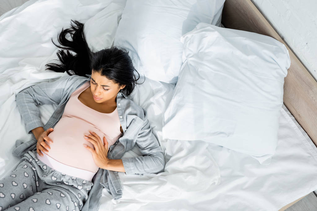 vista superior de la mujer embarazada afroamericana en pijama tocando el vientre y sintiendo dolor
  - Foto, Imagen