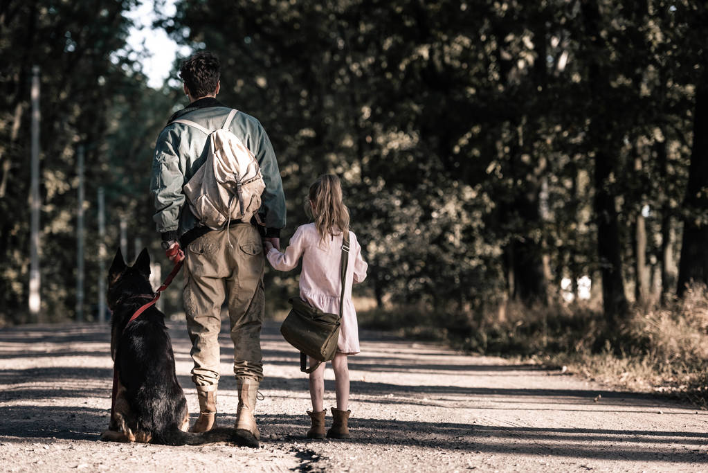 Вид сзади человека, держащего за руки ребенка рядом с немецкой пастушьей собакой, пост-апокалиптическая концепция
 - Фото, изображение