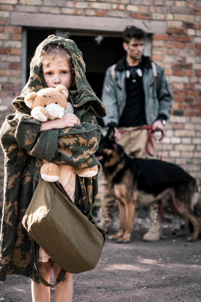 放棄された建物の近くにドイツの羊飼いの犬と男の近くにテディベアを保持する子供の選択的な焦点,黙示録的な概念を投稿 - 写真・画像
