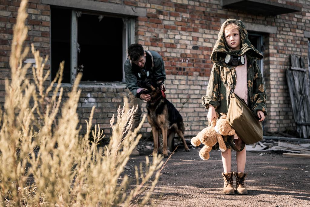 enfoque selectivo de niño sosteniendo oso de peluche cerca de hombre con perro pastor alemán cerca de edificio abandonado, concepto post apocalíptico
 - Foto, Imagen
