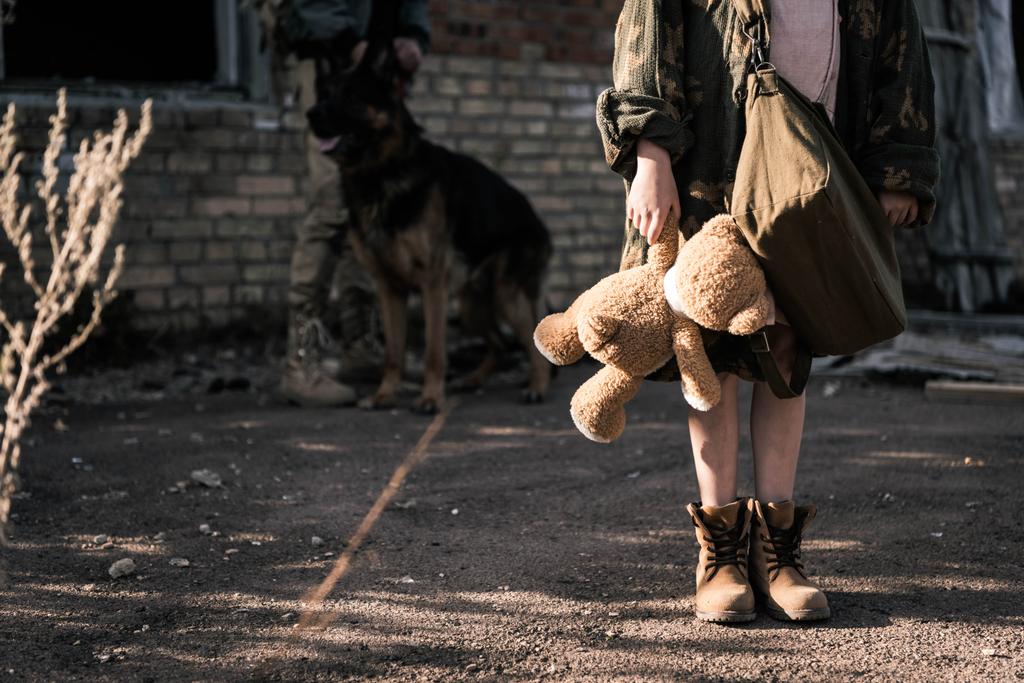 cropped view of kid holding teddy bear near man with german shepherd dog near abandoned building, post apocalyptic concept - Photo, Image