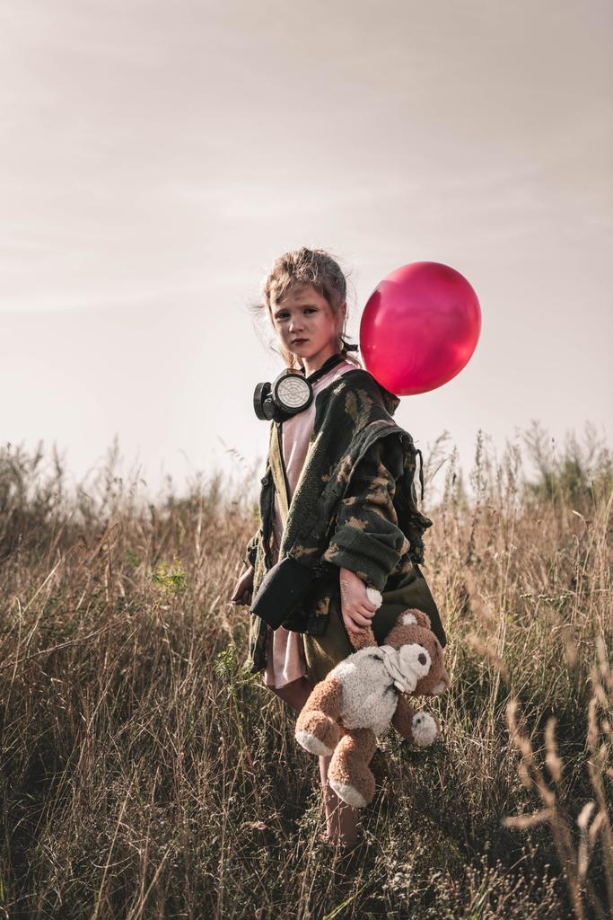 selektivní zaměření roztomilého kluka s plynovou maskou a plyšovým medvídkem držícím balónek, post apokalyptický koncept - Fotografie, Obrázek