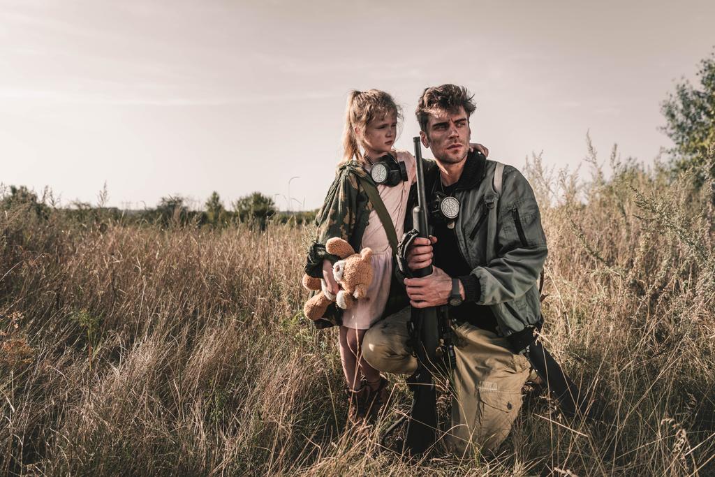 handsome man holding gun near kid with soft toy in field, post apocalyptic concept - Photo, Image