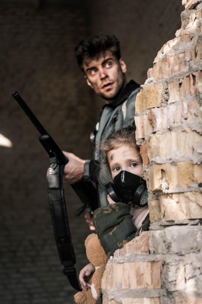 селективное внимание ребенка в противогазе рядом с красивым мужчиной с пистолетом, постапокалиптическая концепция
 - Фото, изображение