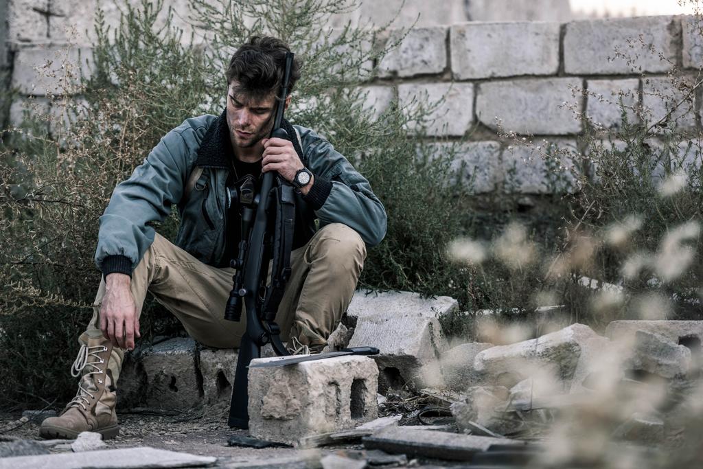 selective focus of handsome man sitting with gun near brick wall outside, post apocalyptic concept - Photo, Image