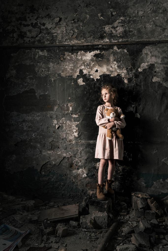 засмучена дитина стоїть і тримає плюшевого ведмедя в брудній кімнаті, пост-апокаліптична концепція
 - Фото, зображення