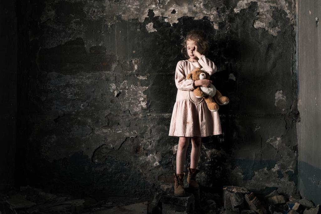 Frustriertes Kind weint, während es Teddybär in schmutzigem Raum hält, postapokalyptisches Konzept - Foto, Bild