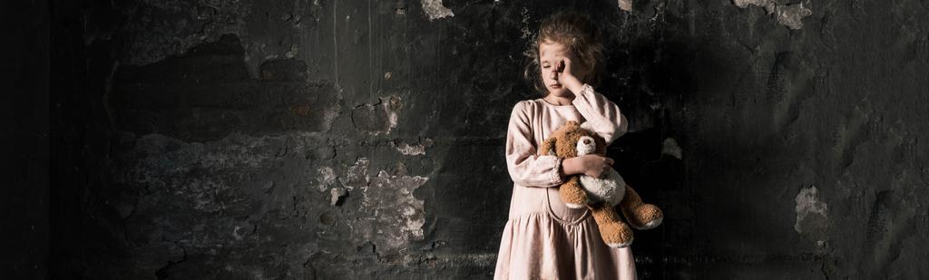 Panoramaaufnahme eines frustrierten Kindes, das weint, während es einen Teddybär in einem schmutzigen Raum hält, postapokalyptisches Konzept - Foto, Bild