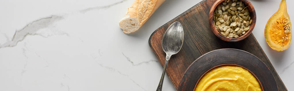 вид сверху на осенний суп из тыквы возле хлеба и семян тыквы на мраморной поверхности, панорамный снимок
 - Фото, изображение