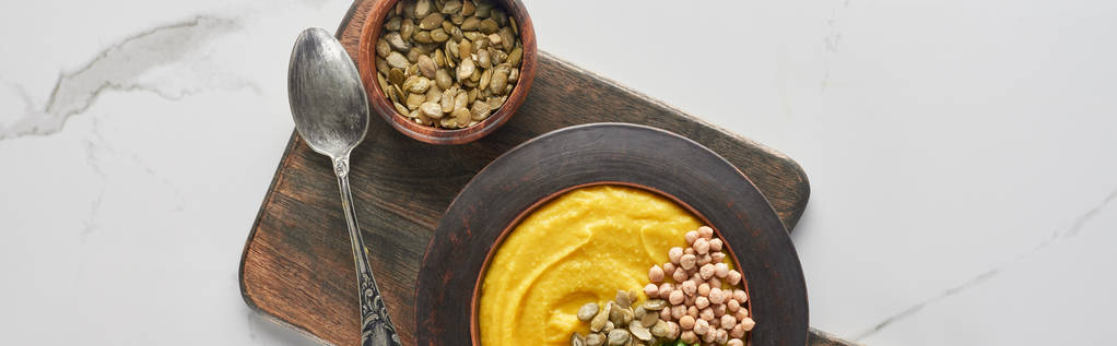 panoramic shot of autumnal mashed pumpkin soup in bowl on wooden cutting board with spoon and seeds on marble surface - Photo, Image
