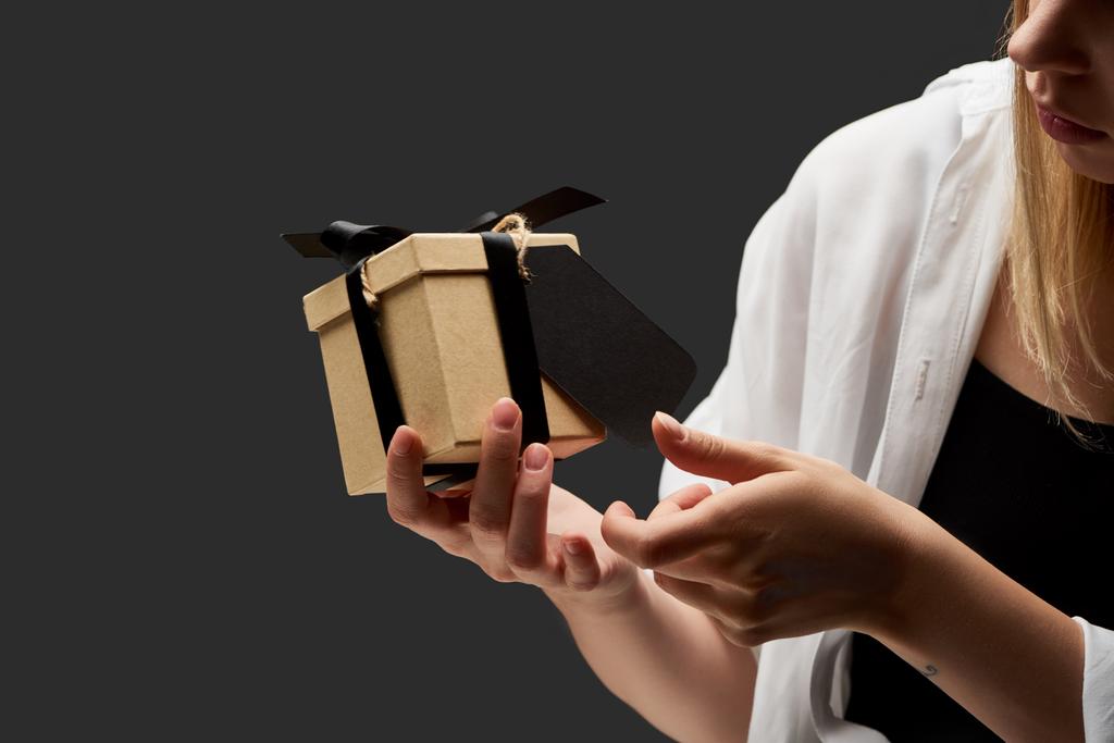 oříznutý pohled na ženu držící dárkovou krabici s černým prázdným štítkem v ruce izolované na černé - Fotografie, Obrázek