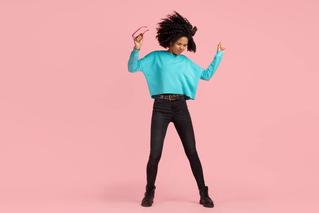 Pleine photo longue de jeune femme afro-américaine excitée avec un sourire éclatant habillé de vêtements décontractés, lunettes et écouteurs dansent sur fond rose
. - Photo, image