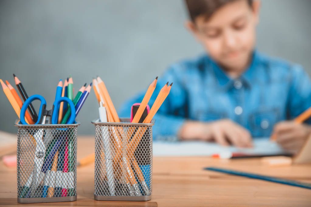 Duas cestas com lápis coloridos, tesouras e marcadores na vanguarda. Estudante escrevendo no fundo embaçado
. - Foto, Imagem