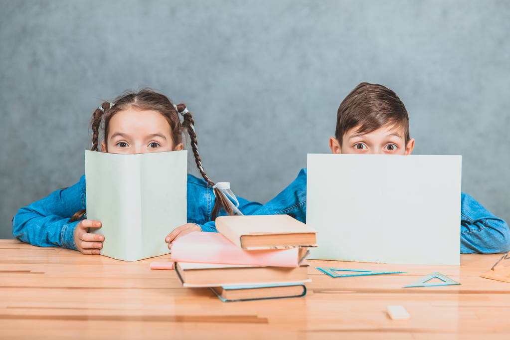 Nieuwsgierige kleine jongen en meisje verbergen achter de vellen papier, kijkend over, op de camera, conspiciuosly. - Foto, afbeelding