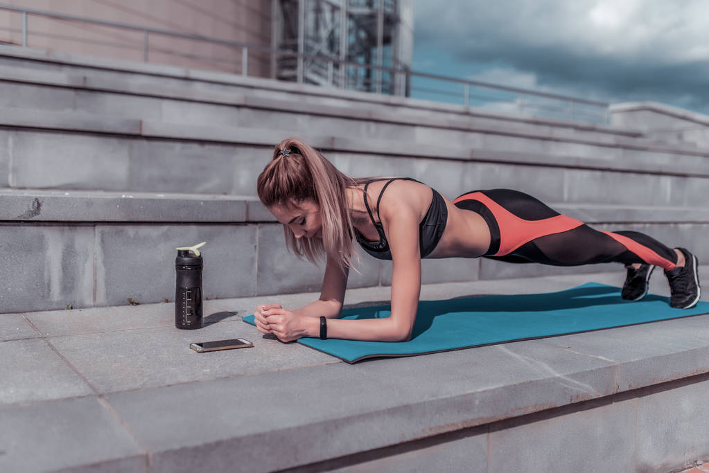 Girl training summer in city, stands emphasis lying down, plank, abs training, abdominal muscles woman back stretching flexibility, smartphone application phone timer, shaker with protein water. - Photo, Image