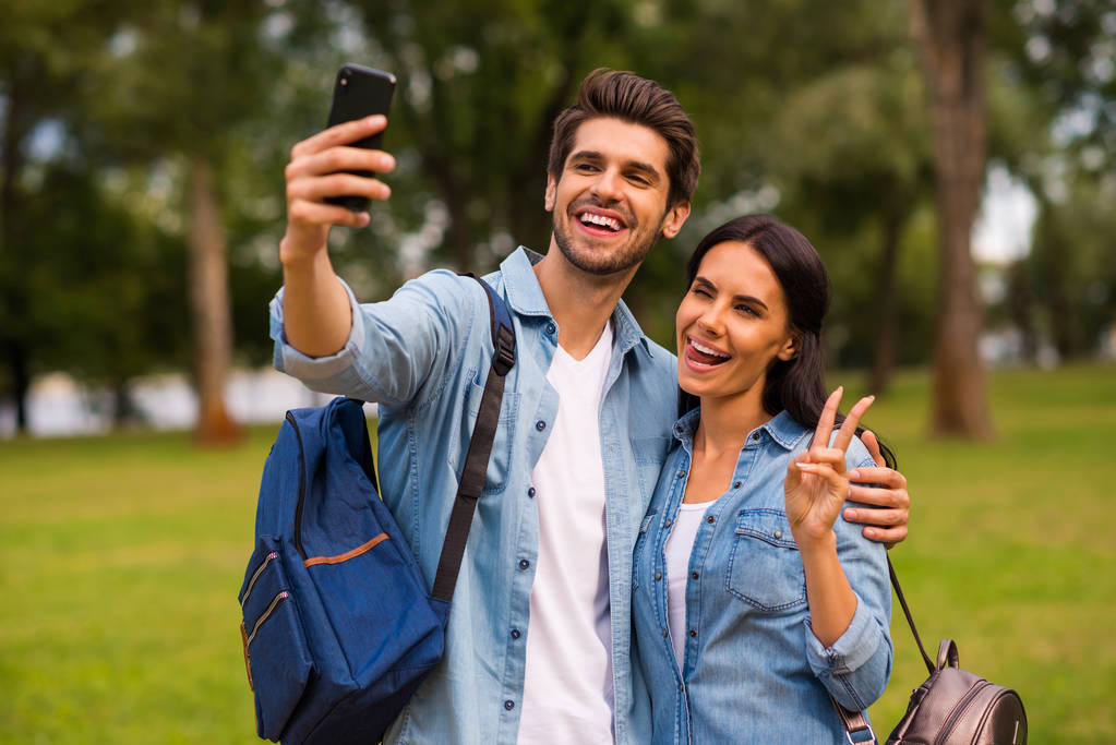 Foto di coppia millenaria utilizzando telefono facendo selfie con simbolo v-segno visita parco verde indossare abbigliamento casual denim
 - Foto, immagini
