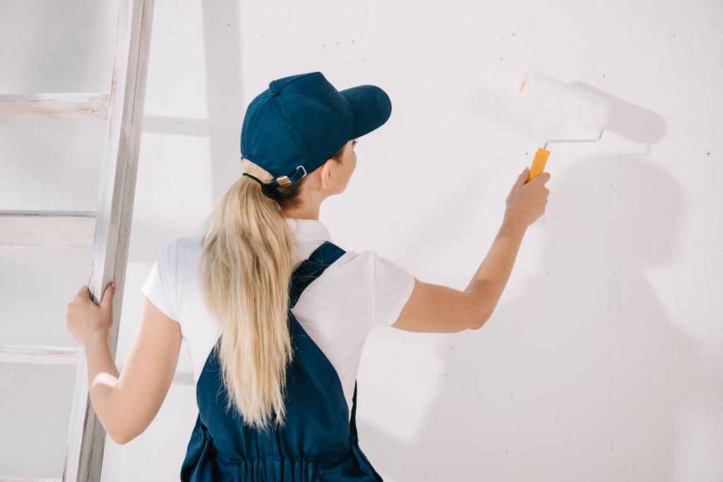 vista trasera del joven pintor en uniforme de pie cerca de la pared blanca y sosteniendo el rodillo de pintura
 - Foto, Imagen
