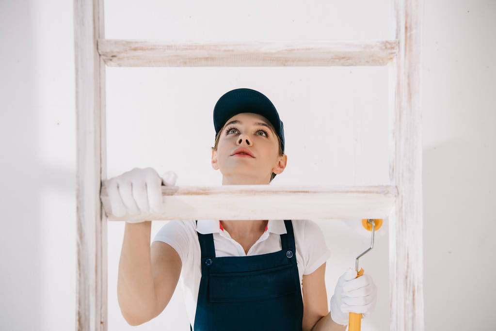 pretty young painter in uniform looking up while climbing on ladder - Photo, Image