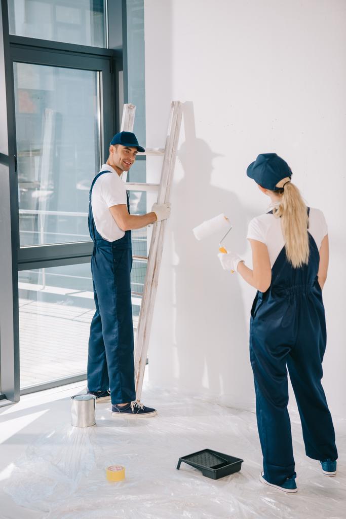 handsome painter standing near ladder while pretty colleague holding paint roller - Photo, Image