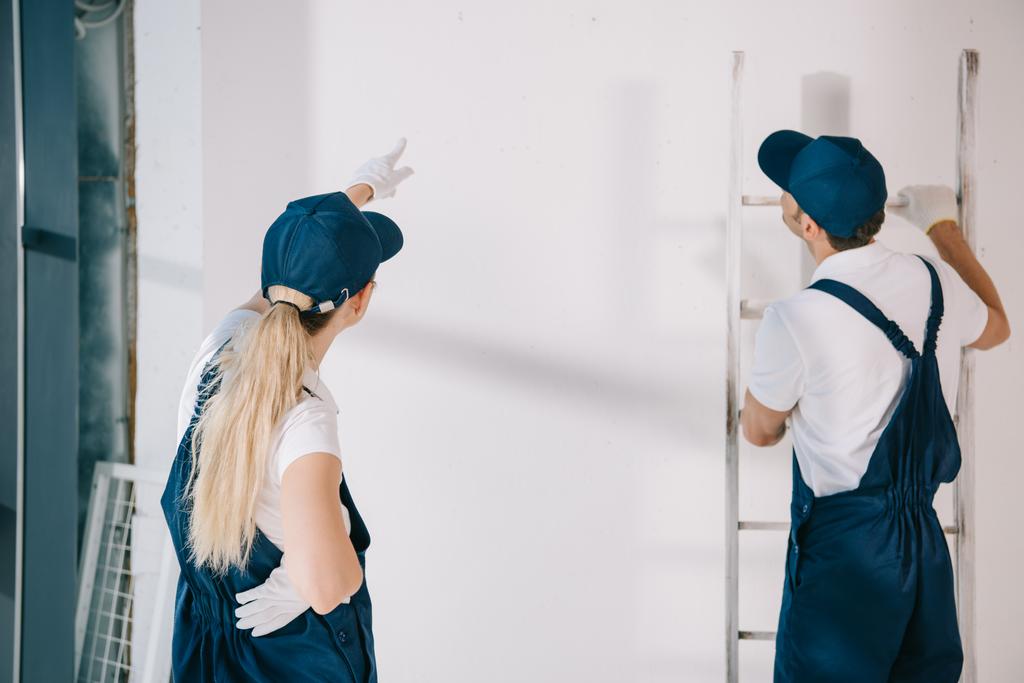 白い壁に指を指差して制服を着た若い画家がはしごを持って同僚の近くに立っている間 - 写真・画像