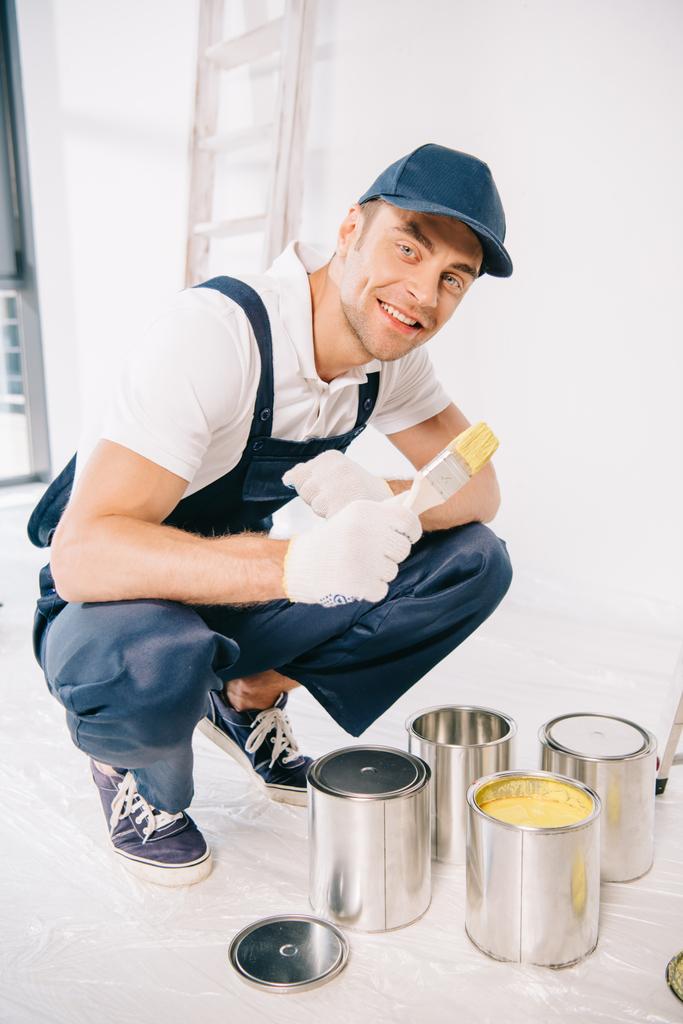 joven pintor guapo en uniforme sosteniendo pincel cerca de latas con pintura y sonriendo a la cámara
 - Foto, imagen