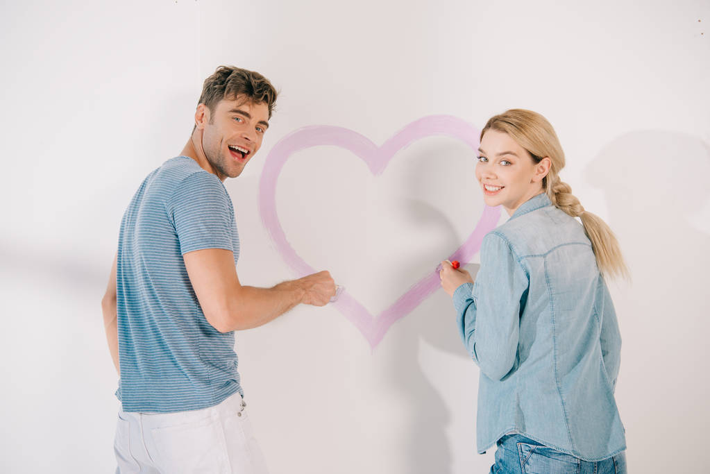glückliches junges Paar lächelt in die Kamera, während es in der Nähe eines rosafarbenen Herzens auf einer weißen Wand steht - Foto, Bild