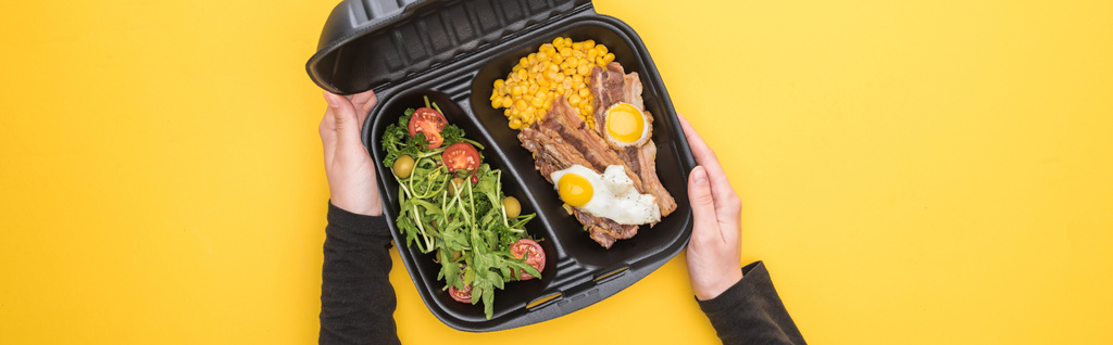 panoramisch schot van de vrouw met eco-pakket met maïs, vlees, gebakken eieren en salade geïsoleerd op geel     - Foto, afbeelding