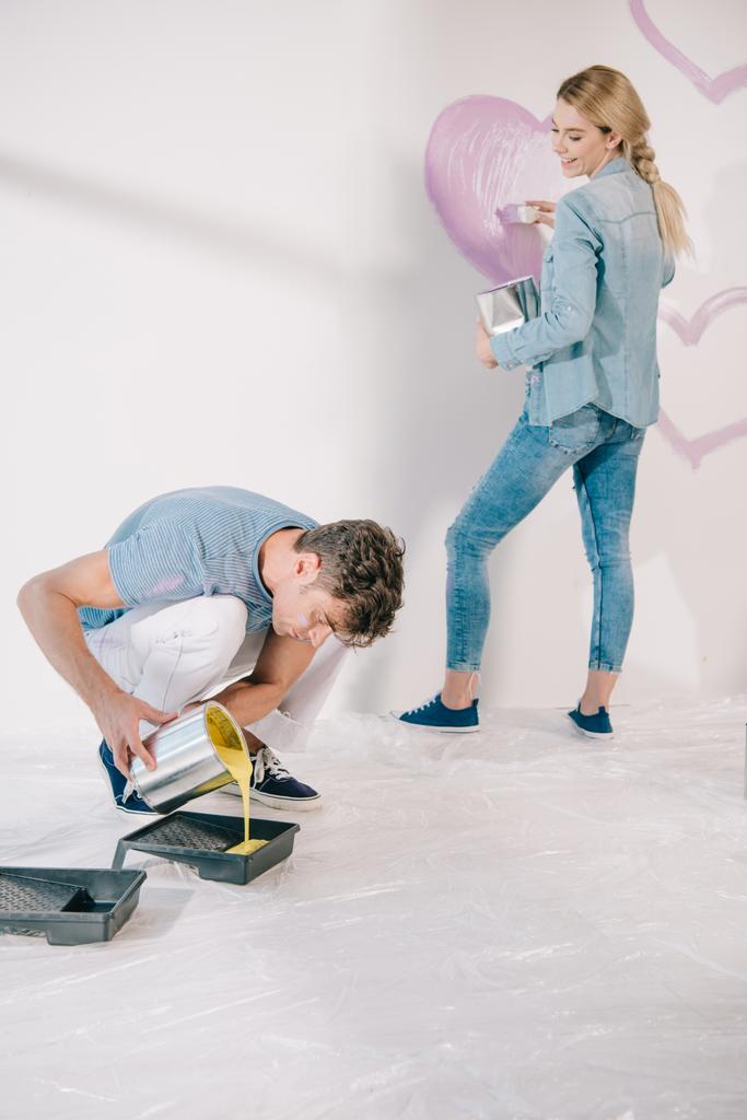 jovem adicionando tinta amarela na bandeja de rolo enquanto namorada desenhando coração rosa na parede branca
 - Foto, Imagem