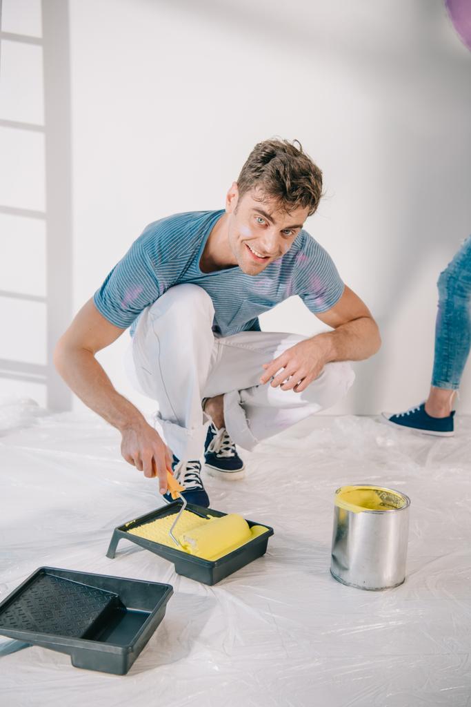 handsome young man putting paint roller into roller tray with yellow paint and smiling at camera - Photo, Image