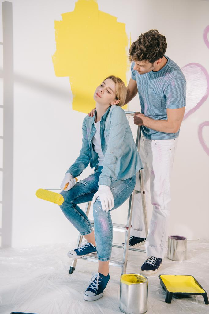 bonito homem de pé perto cansado namorada sentado na escada e segurando amarelo rolo de pintura
 - Foto, Imagem