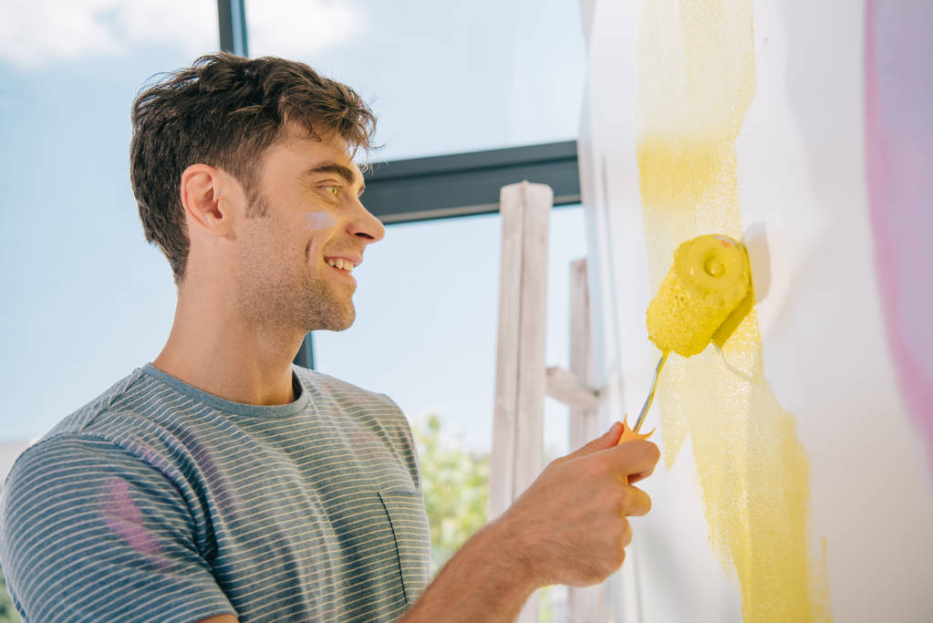 όμορφος νεαρός άνδρας χαμογελά ενώ ζωγραφική τοίχο σε κίτρινο χρώμα με ρολό - Φωτογραφία, εικόνα