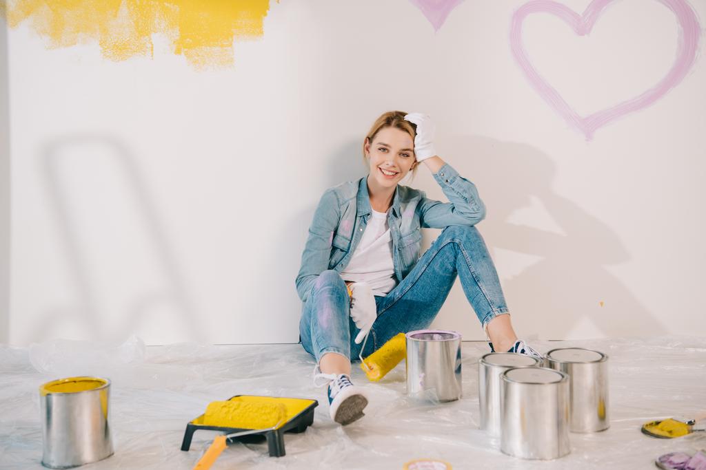 atractiva mujer joven sosteniendo rodillo de pintura amarillo mientras está sentado en el suelo cerca de la pared
 - Foto, imagen