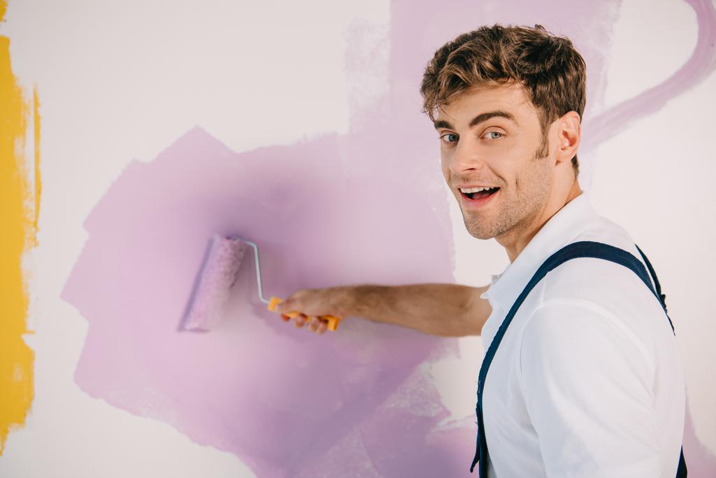 apuesto pintor joven pintura de pared en color rosa con rodillo de pintura y sonriendo a la cámara
 - Foto, imagen