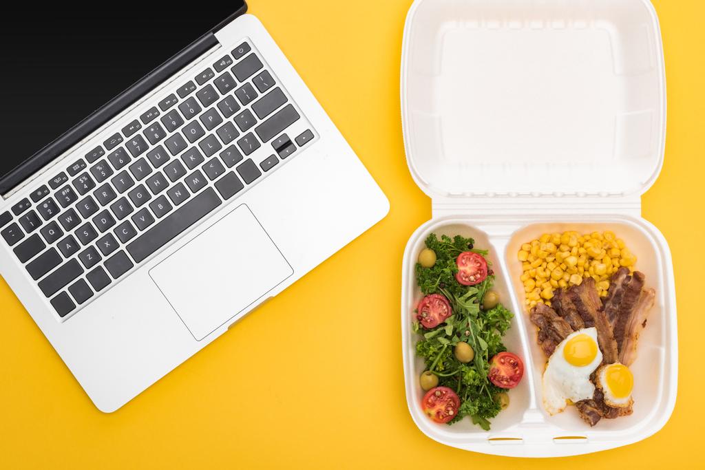 top view of laptop and eco package with corn, meat, fried eggs and salad isolated on yellow  - Photo, Image