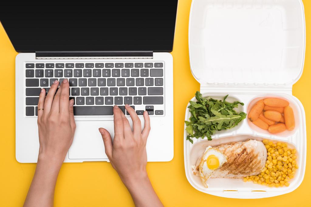 καλλιεργημένη άποψη της γυναίκας που χρησιμοποιεί φορητό υπολογιστή και οικολογική συσκευασία με λαχανικά, κρέας, τηγανητό αυγό και ρόκα απομονώνονται σε κίτρινο  - Φωτογραφία, εικόνα