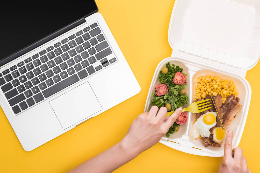 gewas uitzicht op vrouw eten uit eco-pakket met maïs, vlees, gebakken eieren en salade geïsoleerd op geel  - Foto, afbeelding