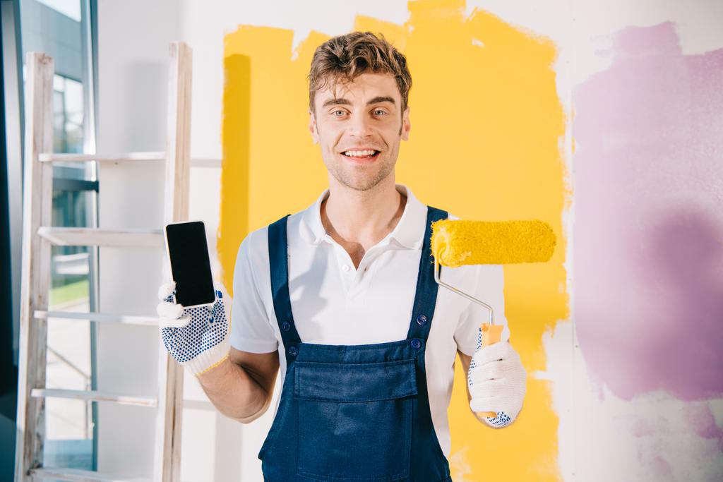 alegre pintor en uniforme sosteniendo teléfono inteligente con pantalla en blanco y rodillo de pintura mientras sonríe a la cámara
 - Foto, Imagen