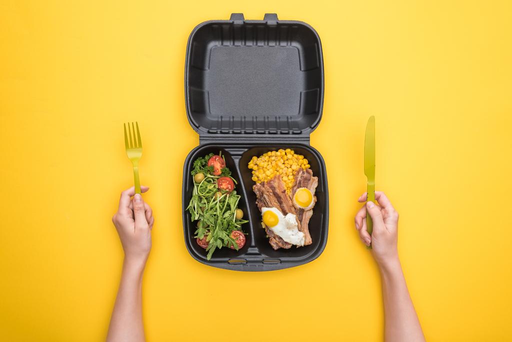vue recadrée d'une femme tenant une fourchette en plastique, un couteau et un emballage écologique avec du maïs, de la viande, des œufs frits et une salade isolés sur du jaune
  - Photo, image