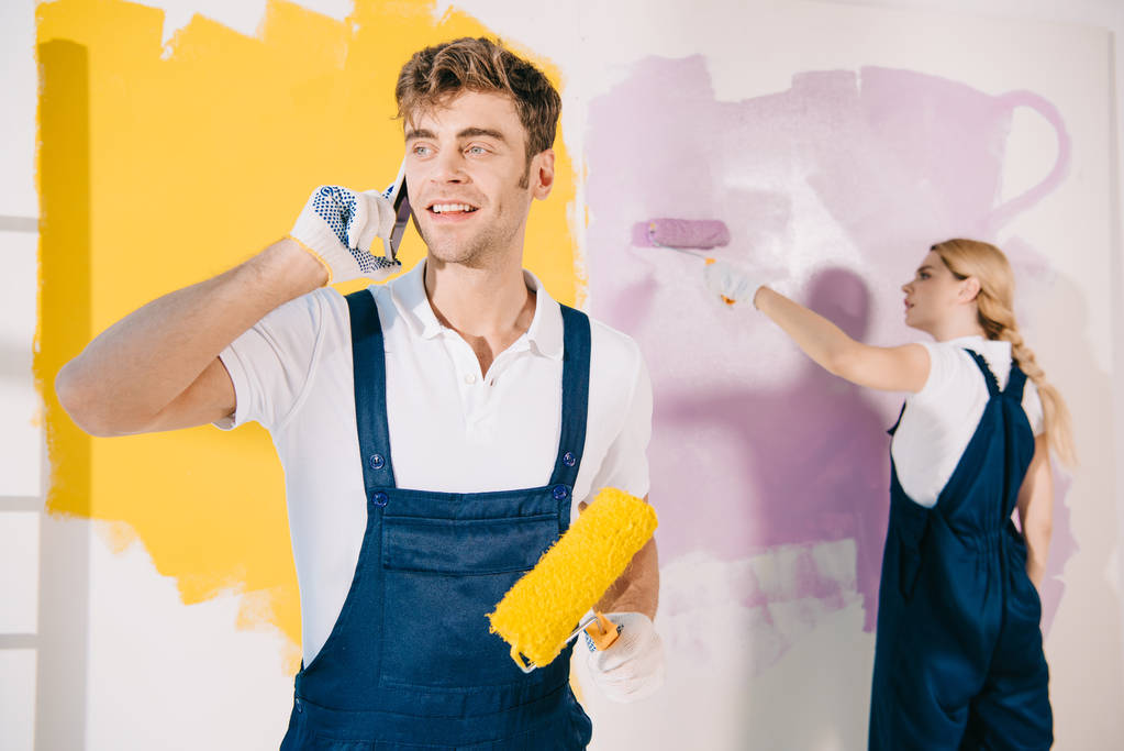 bonito pintor falando no smartphone enquanto atraente colega pintura parede
 - Foto, Imagem