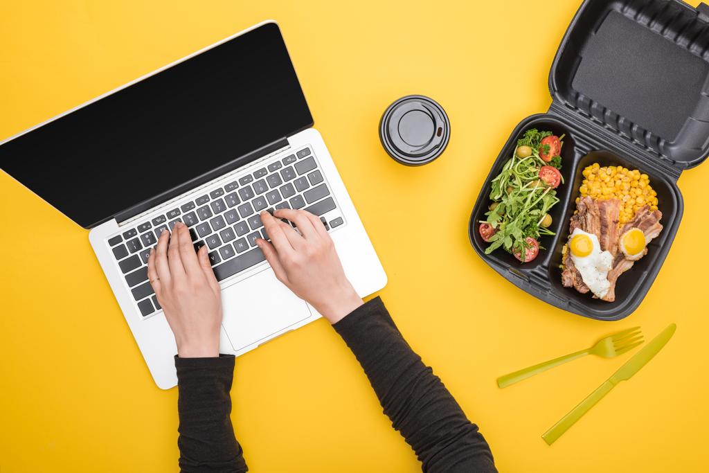 vista recortada de la mujer usando el ordenador portátil y el paquete ecológico con maíz, carne, huevos fritos y ensalada, taza de papel aislado en amarillo
  - Foto, imagen