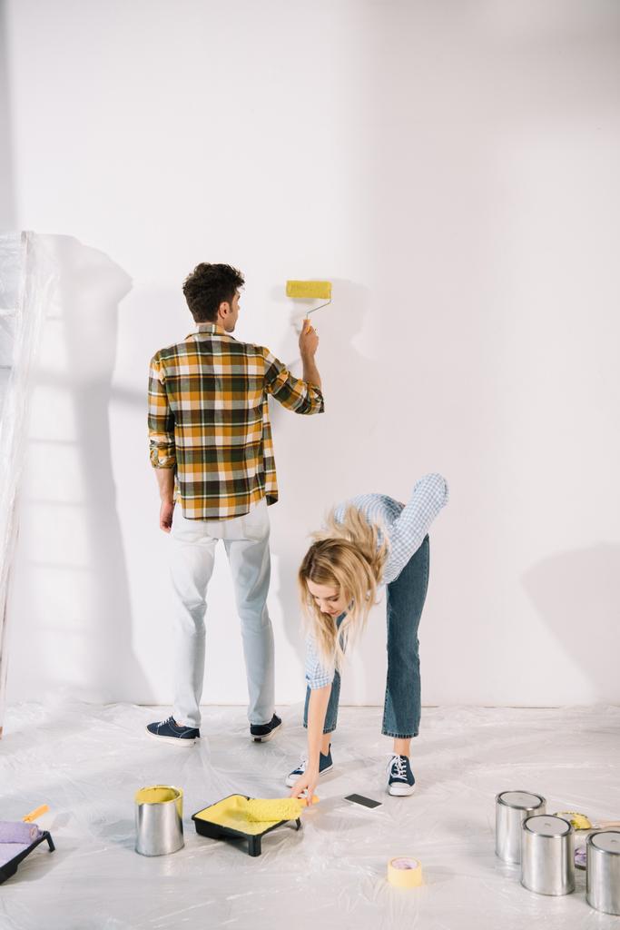 jonge vrouw zetten verf roller in roller lade met gele verf terwijl vriendje schilderen muur - Foto, afbeelding