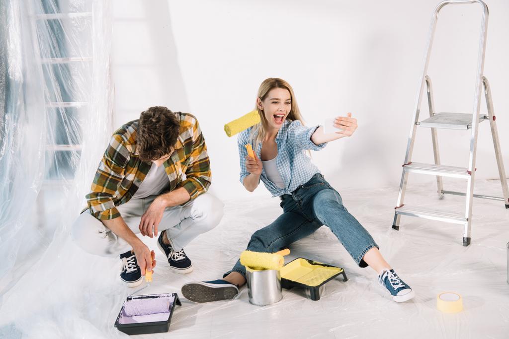 alegre joven mujer sosteniendo amarillo pintura rodillo y tomando selfie mientras novio poner pintura rodillo en rosa pintura
 - Foto, Imagen