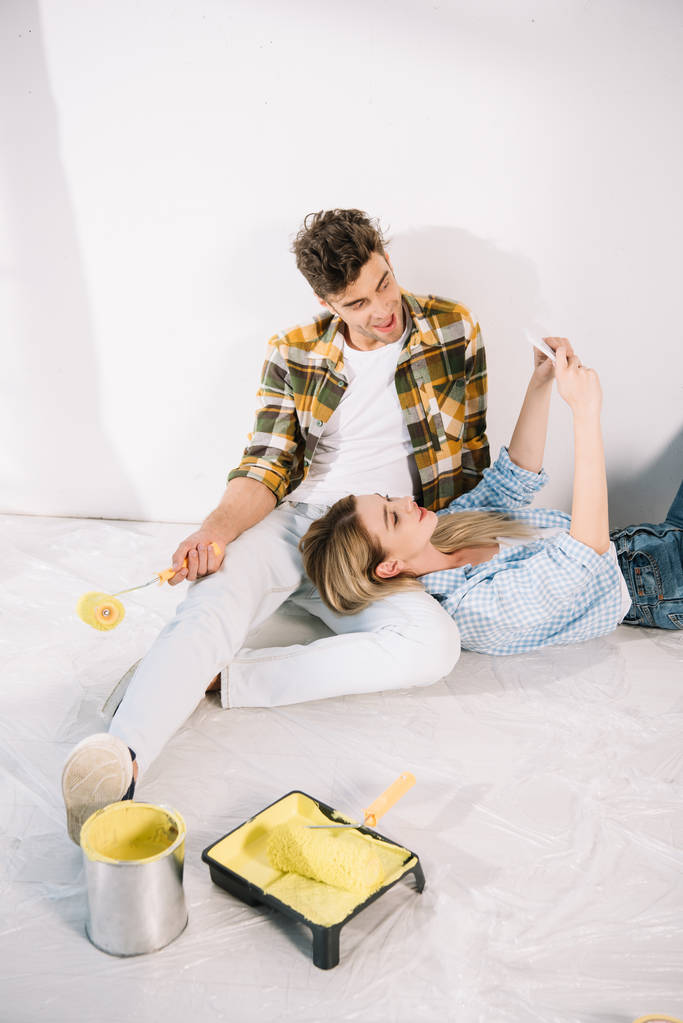 Nuori nainen ottaa selfie poikaystävänsä kanssa tilalla keltainen maalirulla istuessaan lattialla
 - Valokuva, kuva