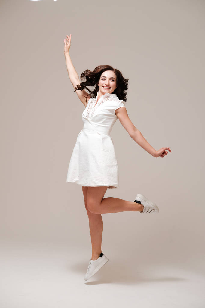 Studioporträt einer lässigen jungen Frau in weißem Kleid und Turnschuhen, die auf schlichtem Hintergrund springt - Foto, Bild