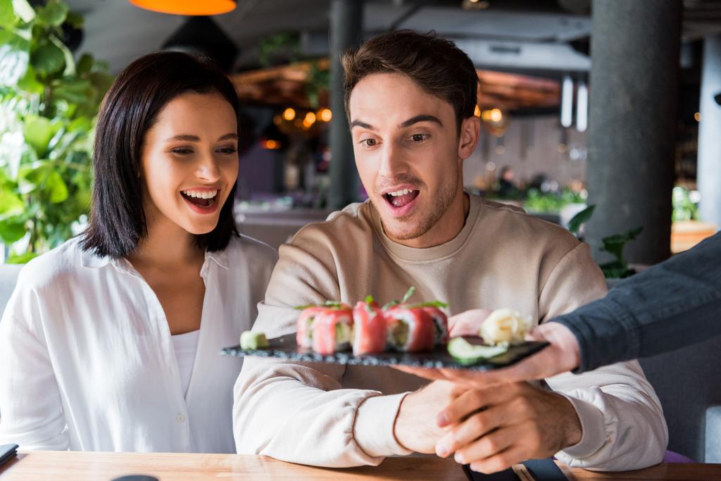vista recortada del camarero sosteniendo el plato con sushi sabroso cerca de la pareja sorprendida en el bar de sushi
  - Foto, imagen