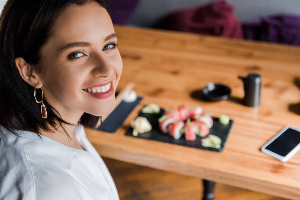 επιλεκτική εστίαση της ευτυχούς γυναίκας κοντά smartphone με λευκή οθόνη και νόστιμο σούσι στο εστιατόριο  - Φωτογραφία, εικόνα