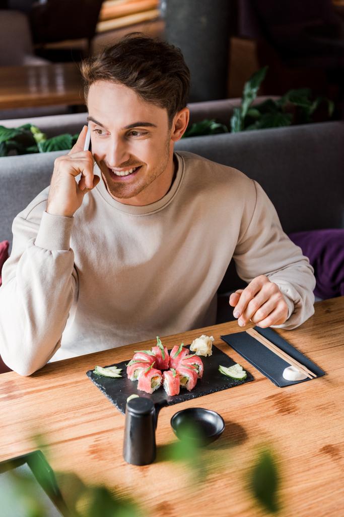Selektiver Fokus des glücklichen Mannes, der in einer Sushi-Bar in der Nähe schmackhafter Mahlzeiten mit dem Smartphone spricht  - Foto, Bild