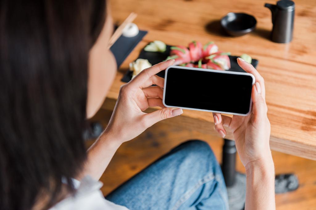 foco seletivo da mulher segurando smartphone com tela em branco perto de sushi no restaurante
  - Foto, Imagem